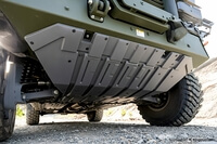 The image shows the underside of a military vehicle, highlighting its protective underbody plating and suspension components.